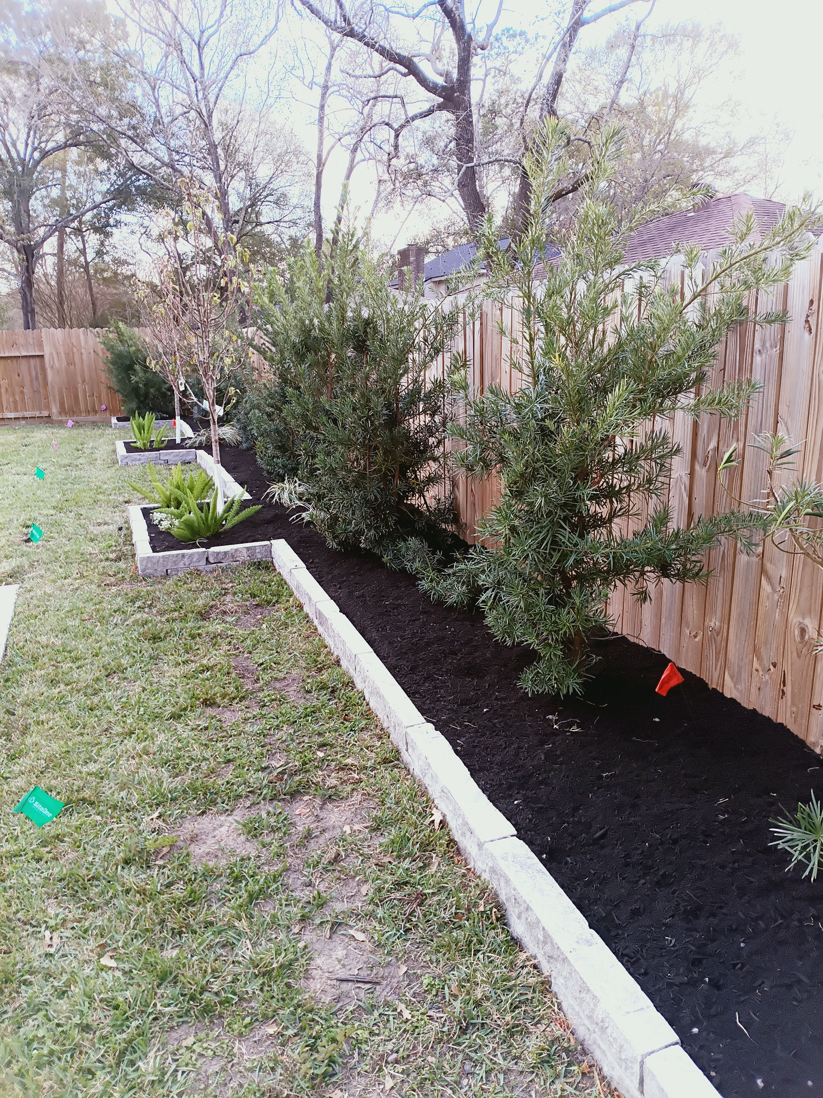 Long planting bed with dark mulch, shrubs and a light stone border beside a wooden fence