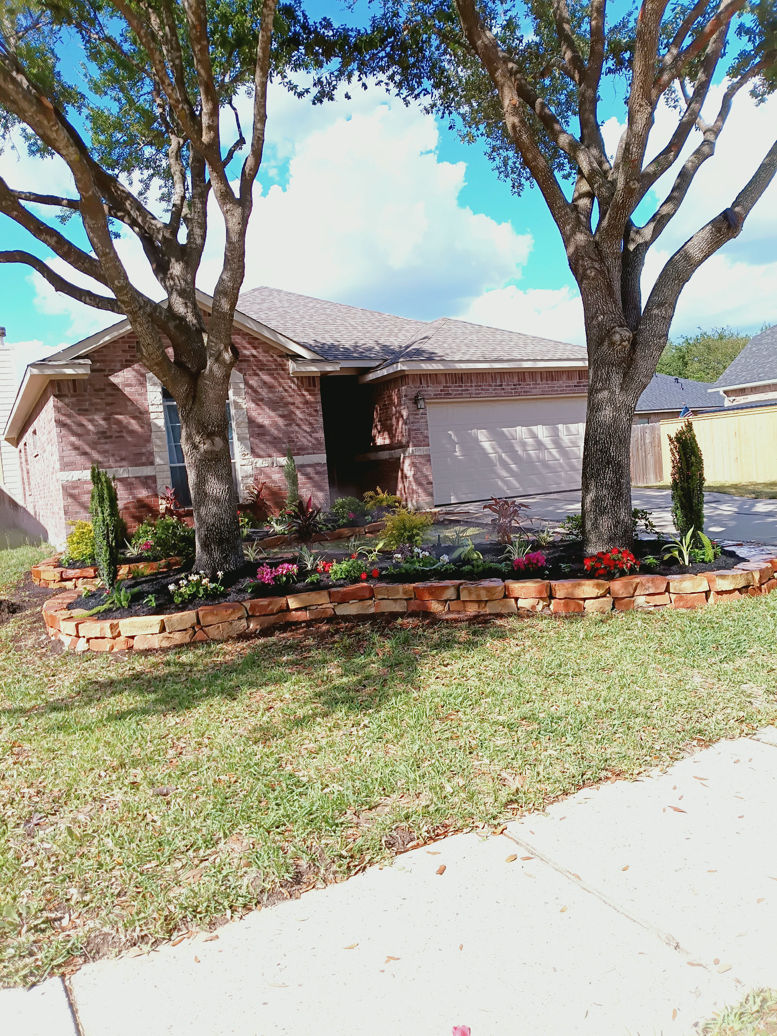 Stone-edged front garden with colorful flowers and planting beds around two mature trees beside a brick home